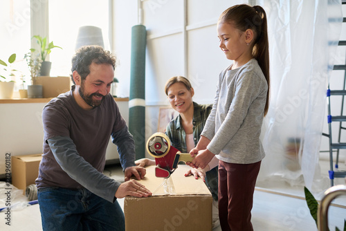 Adorable little child sealing packed cardboard box with adhesive tape while helping her father and mother during relocation