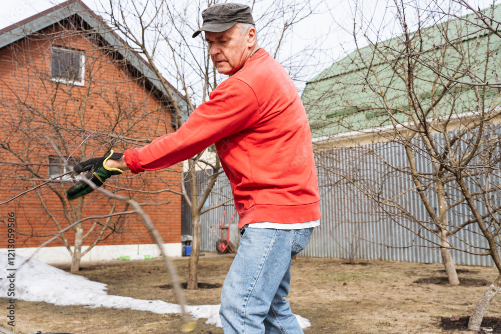 A man in a red sweatshirt and black cap is pruning tree branches in a garden. The scene captures outdoor work in early spring, with bare trees and a rural setting in the background.