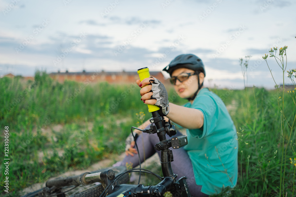 Woman wearing a helmet and sunglasses sitting in a field. She is resting beside her mountain bike. The womans calm demeanor suggests she is taking a break from her bike ride. Woman wearing a helmet and sunglasses sitting in a field. She is resting beside her mountain bike. The womans calm demeanor suggests she is taking a break from her bike ride.