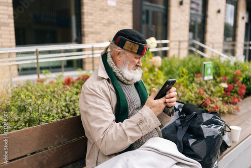 Side view of aged beggar man wrapped in blanket using smartphone sitting on urban bench outside building on cold autumn day. Concept of poverty and vulnerability, evoking empathy and compassion. photo