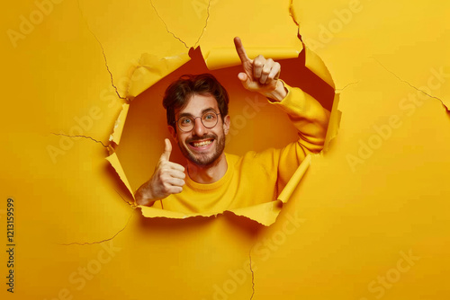 Obraz na płótnie A man in a yellow sweater peeks through a torn paper wall, thumbs up, smiling. Concept of breakthrough and positivity. For creativity and energy. w salonie