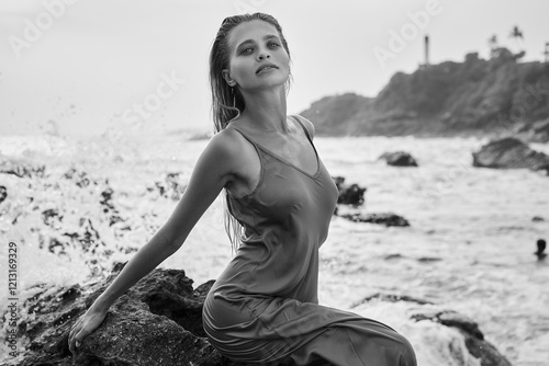 Young woman in wet dress by ocean rocks, feels free. Her look is serene, captures divine beauty. Waves crash, nature vibes, femininity, elegance. Black and white style, timeless allure, summer day. photo