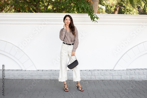 Young beautiful brunette woman wearing striped brown shirt, white denim pants, black heeled sandals and small handbag standing outdoor on a city street. Trendy casual outfit. Street fashion.