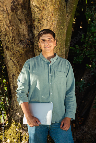 Smiling student holding laptop leaning against tree in park photo