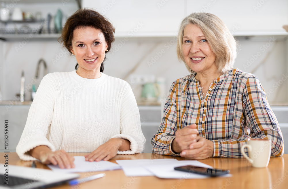 Smiling old and middle-aged women posing at the kitchen-table with sheets of paper and laptop in front of them Smiling old and middle-aged women posing at the kitchen-table with sheets of paper and laptop in front of them