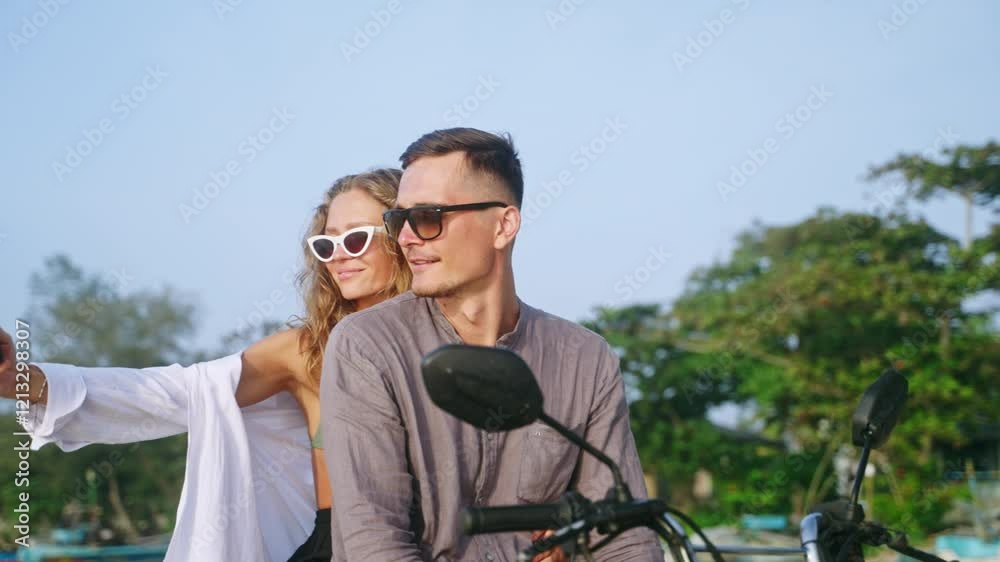 Young romantic couple sit on motorcycle on sea beach at sunset. Female travel blogger filming vlog, taking selfie photo on smartphone. Man, woman in love relax on motorbike trip to ocean coast.