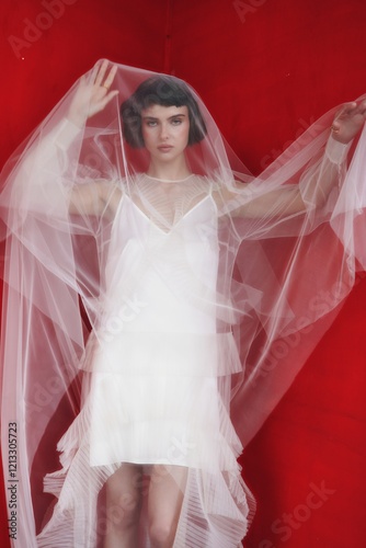 Model poses in a flowing white dress with translucent fabric against a vibrant red background during an artistic fashion shoot