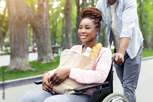 Millennial black handicapped woman in wheelchair holding paper bag with food, going shopping with her boyfriend. Young married disabled couple passing city park on way home from store, panorama