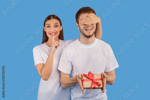 It's A Secret. Girl covering her boyfriend eyes, making surprise and silence gesture, isolated over pastel purple studio wall