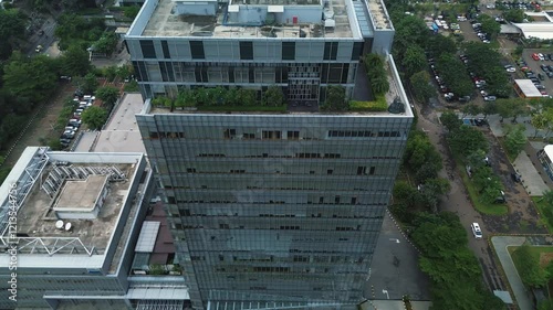 Drone footage of a tall modern office building, surrounded by parking lots and lush green trees.  High-rise cityscape view.