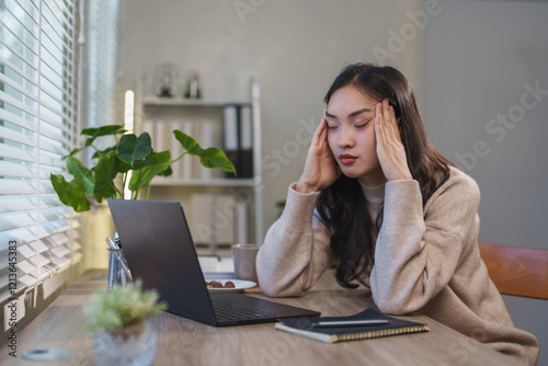 Young asian businesswoman having a headache and massaging her temples while working on a laptop from her home office, showing the challenges of remote work and the importance of mental health