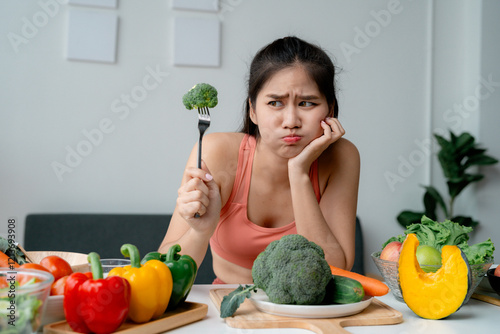 Asian sportswoman looking with disgust at broccoli on a fork while sitting at a table full of vegetables