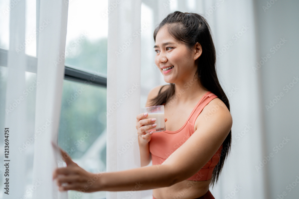 Happy athletic woman enjoying protein shake after workout, opening curtains and looking out the window in her modern apartment