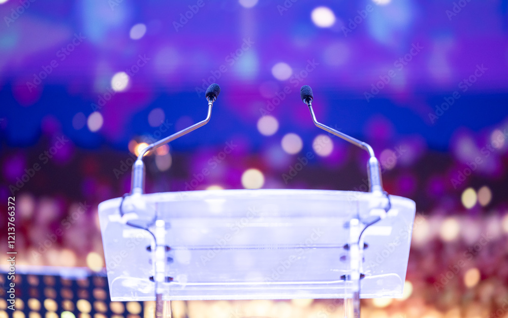 Two microphones on a clear podium with colorful bokeh lights in the background. Perfect for events and public speaking themes. Speaker desk.
