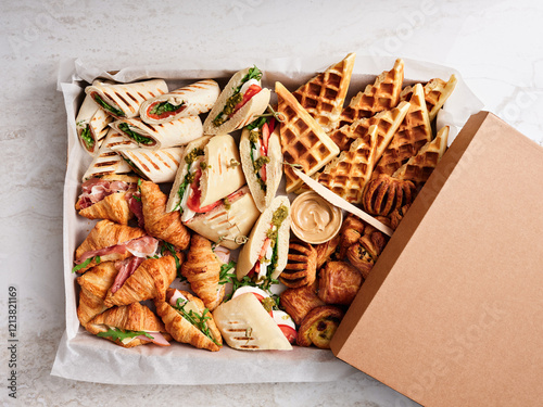 A delightful spread features fresh croissants, gourmet sandwiches filled with meats and vegetables, and golden waffles served with a creamy dipping sauce, perfect for a morning gathering