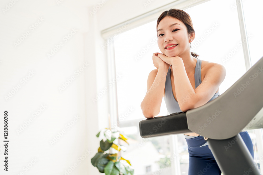 A young attractive active Asian woman smiles while resting on a treadmill in a bright, modern gym space with natural light and greenery. Indoor working out or exercise at home, wearing sportswear. A young attractive active Asian woman smiles while resting on a treadmill in a bright, modern gym space with natural light and greenery. Indoor working out or exercise at home, wearing sportswear.