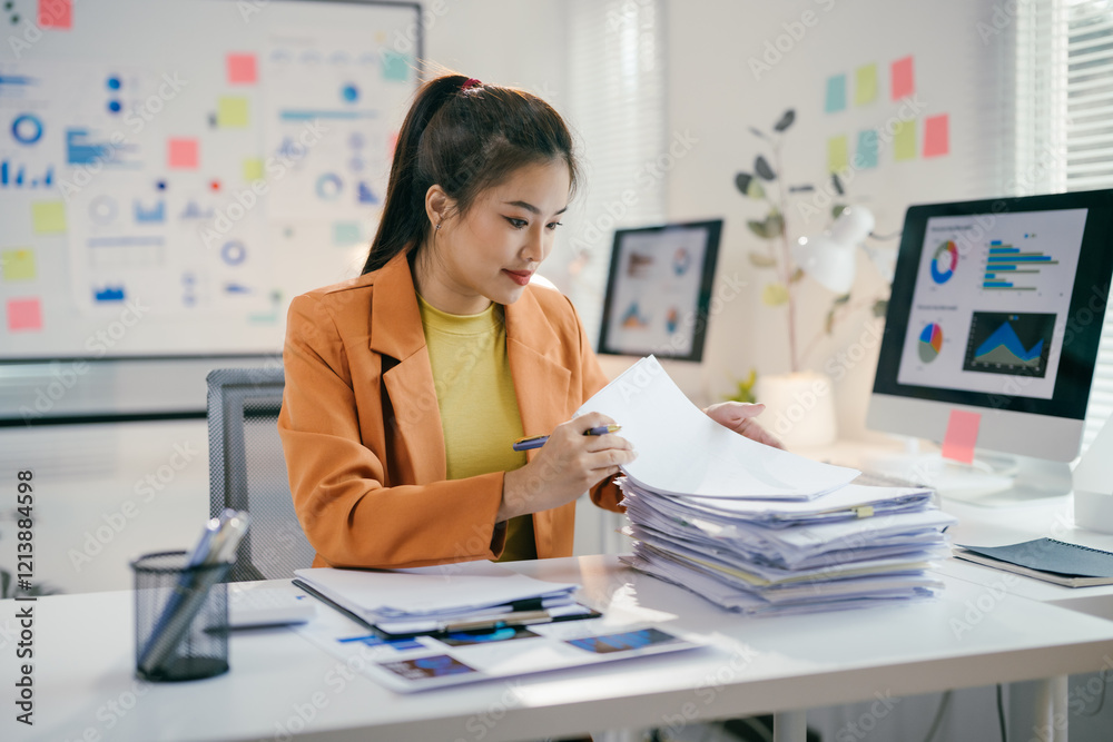 Asian businesswoman reviewing paperwork and using computer with data analysis displayed on screen Asian businesswoman reviewing paperwork and using computer with data analysis displayed on screen