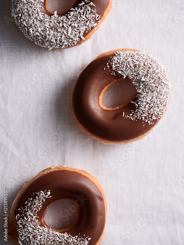 Three chocolate-glazed donuts are displayed on a white cloth, two topped with coconut flakes. The light creates a tempting ambiance around the treats, perfect for dessert lovers photo