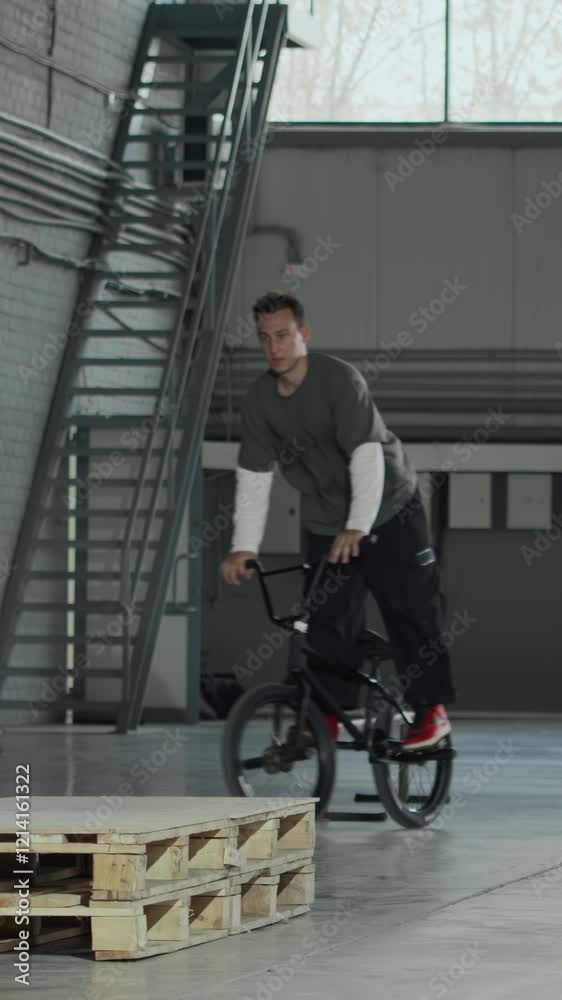 Vertical full stab shot of young Caucasian guy stunt riding on BMX bicycle in hangar and failing to do clean spin, practicing with friend Vertical full stab shot of young Caucasian guy stunt riding on BMX bicycle in hangar and failing to do clean spin, practicing with friend