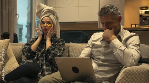 A beautiful woman with a towel on her head and under-eye patches keeps doing her beauty procedures while her boyfriend is working on laptop. photo