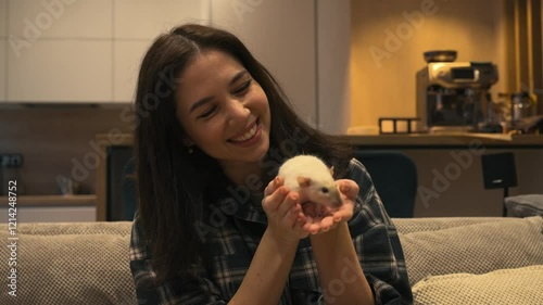 Young brunette woman in a plaid shirt is playing with her home white rat in her hands sitting on the couch, enjoying the interaction photo