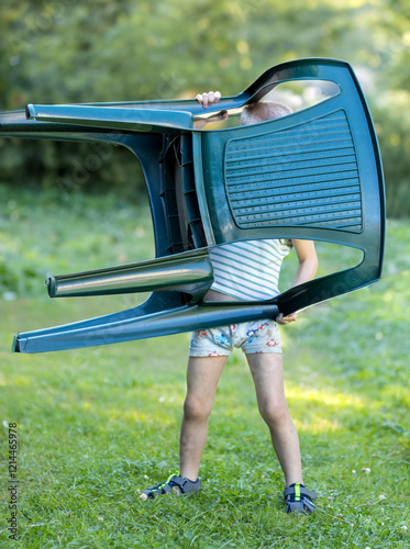 A little boy carries a plastic chair in the garden