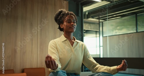 A relaxed girl with black skin in business clothes sits with her eyes closed in the lotus position and meditates on a table in the office