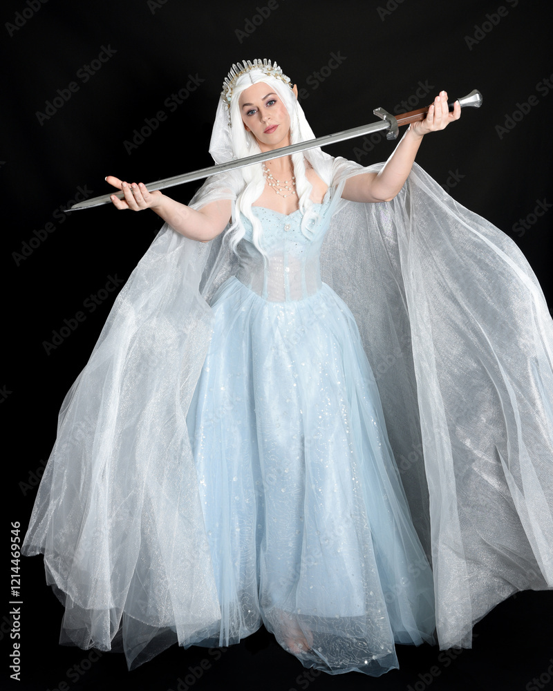 Fototapeta premium full length portrait beautiful blonde fantasy maiden faery wearing medieval fairytale ballgown costume, flowing white hooded cape. standing pose holding a sword weapon, isolated dark studio background