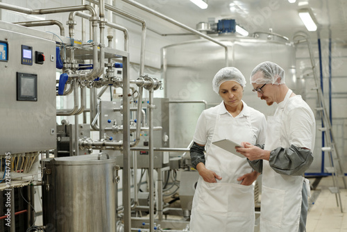 Two factory workers standing in large industrial facility, wearing white uniforms and hairnets, looking at a tablet. Machinery and stainless steel tanks in background