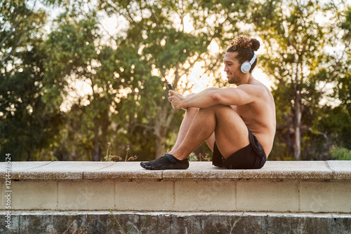 Shirtless athlete relaxing outdoors with headphones and smartphone