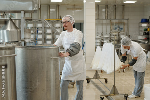 Two workers in an industrial kitchen wearing hairnets and aprons are involved in cheese production. One person is stirring a large vat while another is tending to cheese cloths