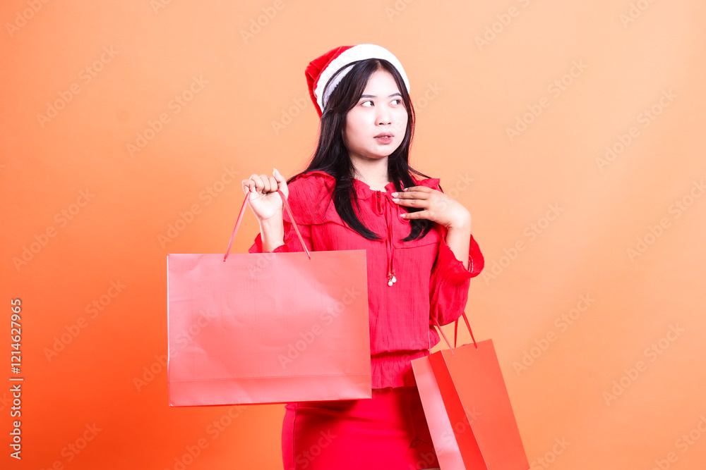 Fototapeta premium portrait of young girl wearing red dress and santa hat, standing sideways left frowning to the left hand holding red bag of christmas gifts and chest, isolated on orange background