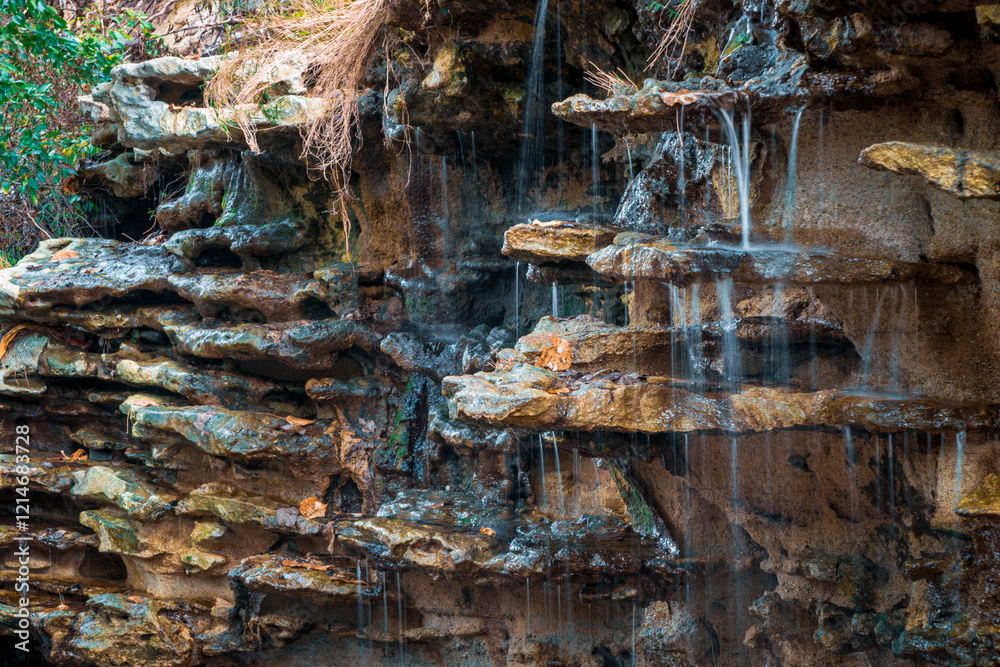 A tranquil waterfall gently flowing over rugged, layered rock formations. The interplay of water and stone creates a soothing atmosphere, enhanced by the lush greenery surrounding the scene