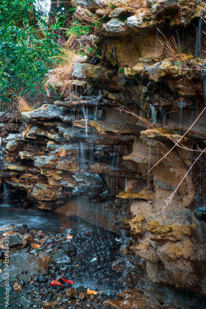 Fototapeta premium A tranquil waterfall gently flowing over rugged, layered rock formations. The interplay of water and stone creates a soothing atmosphere, enhanced by the lush greenery surrounding the scene