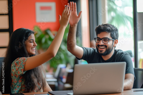 Smiling coworkers celebrating success with a high-five while working on a project in a vibrant, modern office with a laptop on the desk