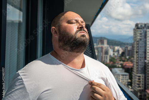 Tired overweight man stands on balcony, fanning shirt collar toward himself. Plump male struggles with hot weather, heat and excessive sweating, discomfort under sun, physical strain, summer challenge