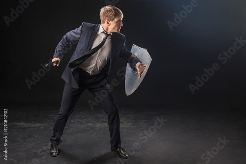 In a dramatic studio pose, a man in a navy suit stands poised with a sword and shield. The dark background emphasizes the contrast between corporate and medieval imagery.