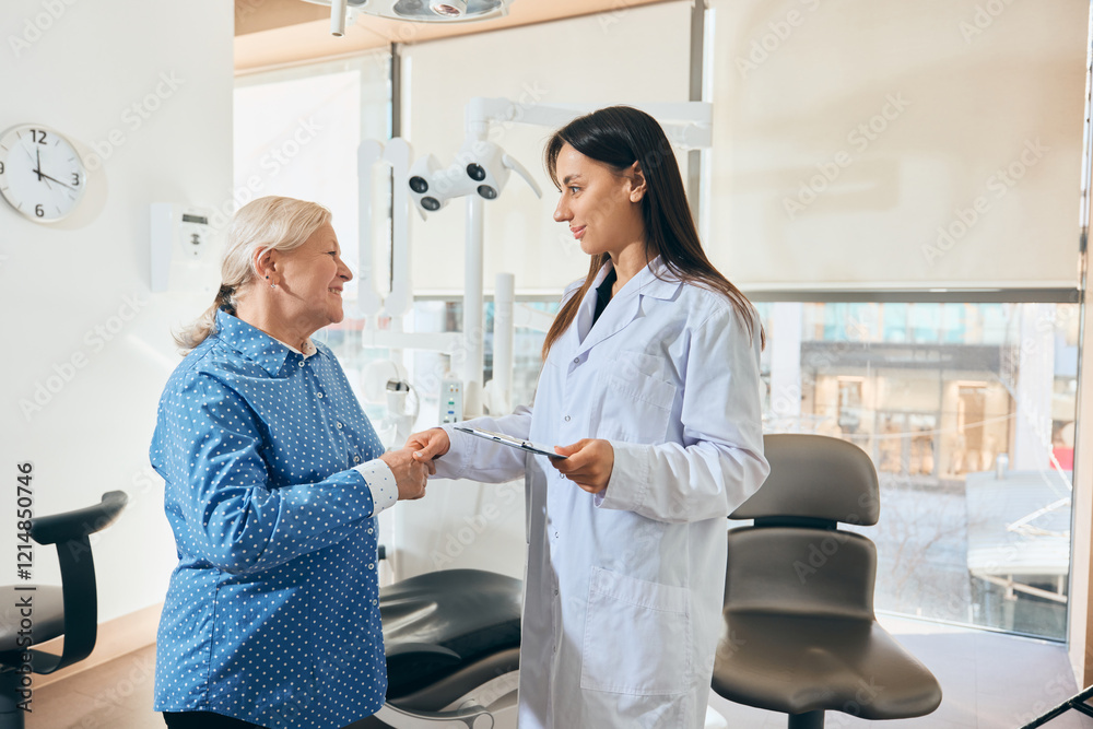 Fototapeta premium Elderly woman shaking hands with female doctor holding clipboard, smiling, dressed in lab coat, both standing in bright medical office. Concept of healthcare and pensioners, medicine.