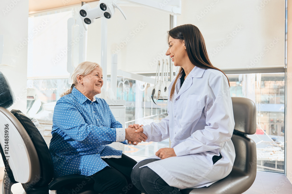 Female doctor seated, shaking hands with smiling elderly woman in blue shirt seated in dental chair, dental equipment in bright office against white background. Concept of healthcare and pensioners. Female doctor seated, shaking hands with smiling elderly woman in blue shirt seated in dental chair, dental equipment in bright office against white background. Concept of healthcare and pensioners.