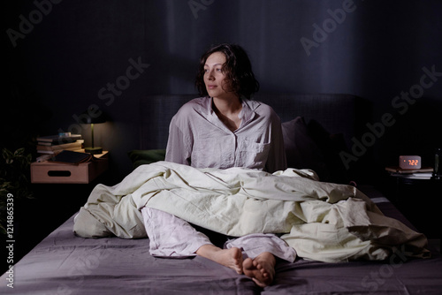 Smiling refreshed woman in comfy pajamas sitting in bed with olive blanket on legs and enjoying illuming beam of sunlight photo