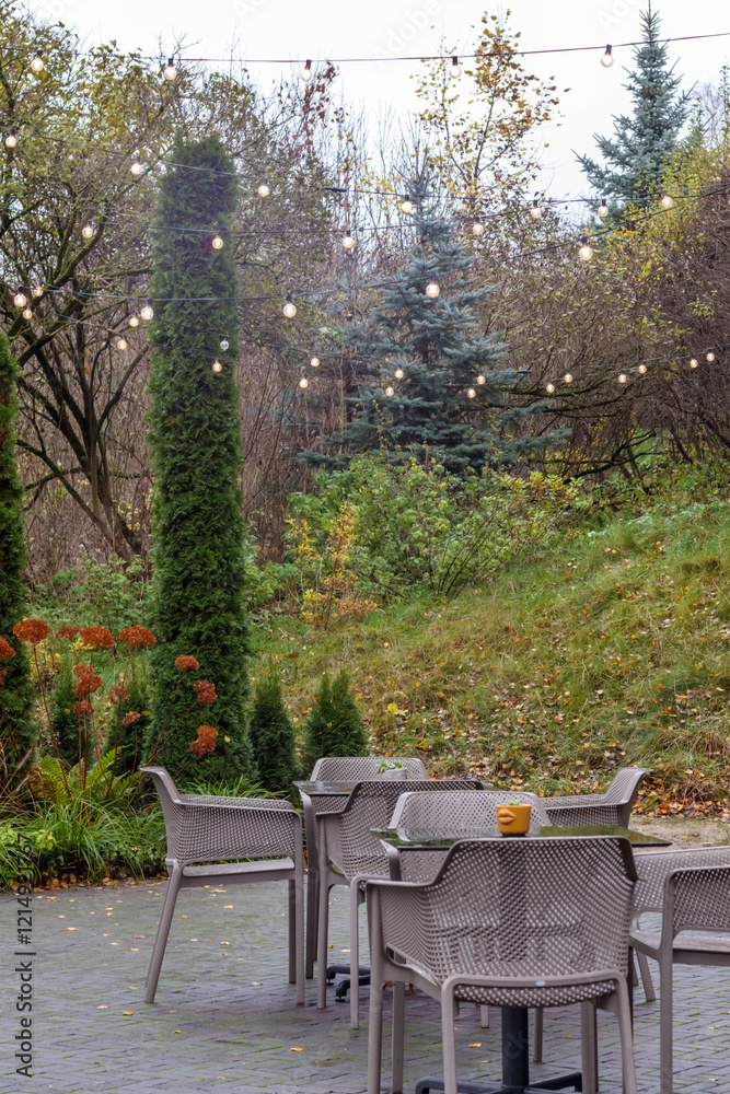 An empty cafe square with a few tables and chairs set in a green park with decorative trees and a string of lights stretched overhead.
An empty cafe square with a few tables and chairs set in a green park with decorative trees and a string of lights stretched overhead.