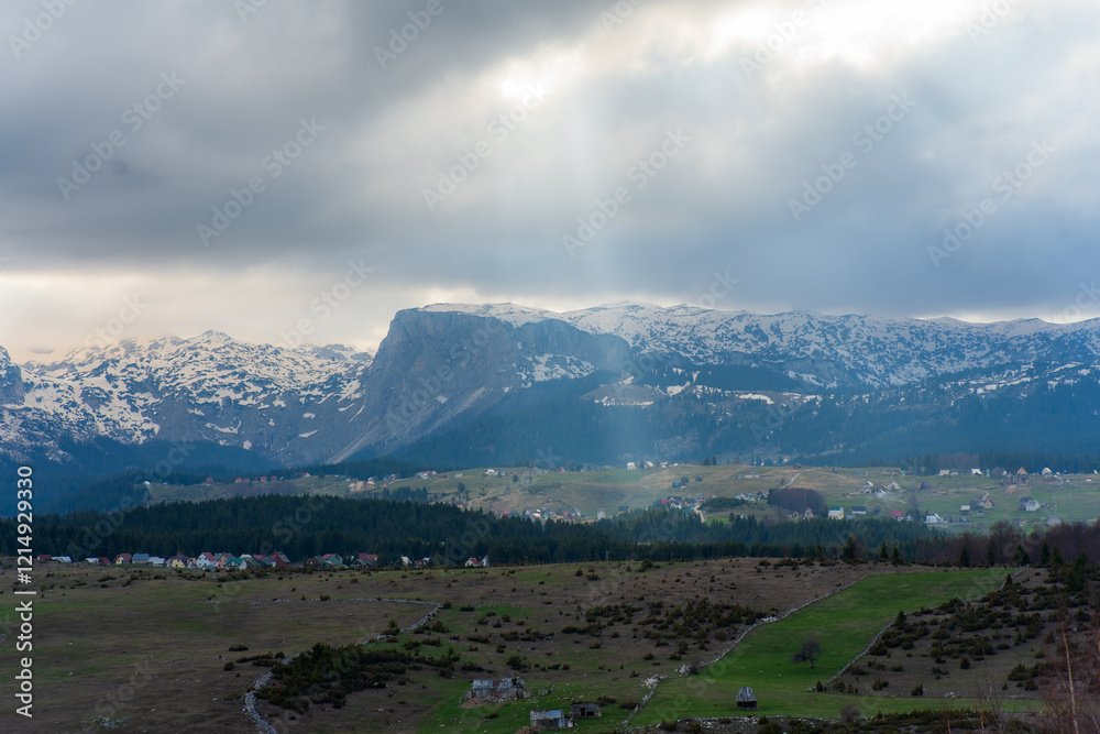 Fototapeta premium Sun rays are illuminating snowy mountain peaks over a green valley with scattered houses and coniferous trees, creating a picturesque landscape in montenegro