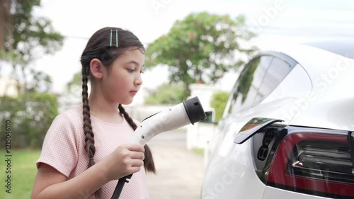 Happy little young girl learn about eco-friendly and energy sustainability as she recharge electric vehicle from home EV charging station. EV car and sustainable future generation concept. Fastidious photo