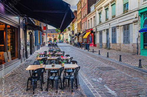 Fototapeta Naklejka Na Ścianę i Meble -  Street restaurant with tables and chairs on narrow cobblestone street, empty road with paving stone, old buildings in Lille city historical center, French Flanders, Hauts-de-France Region, France