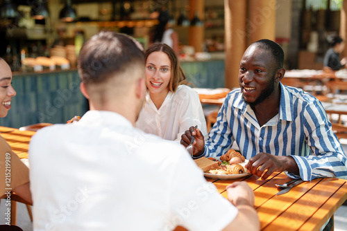 A diverse group of friends, including men and women, share a leisurely breakfast at an outdoor cafe. They are smiling and wearing casual attire, enjoying each other's company on a sunny day.