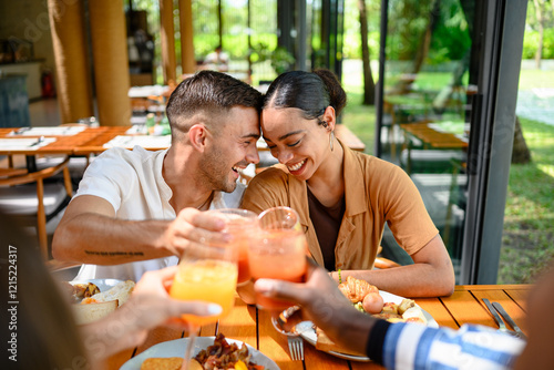 A Caucasian male and multiracial female couple enjoy brunch with diverse friends in a cozy bar, laughing and toasting with fresh juice. The cafe’s bright, sunlit interior offers a warm atmosphere.