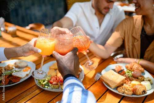 Diverse group of friends joyfully toasting with colorful fresh juice, enjoying a nutritious breakfast together. Plates filled with fruits, pastries, and vegetables create a warm, inviting atmosphere.