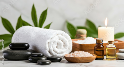 A spa setup with natural elements including a rolled white towel, smooth black massage stones, a wooden tray holding sea salt