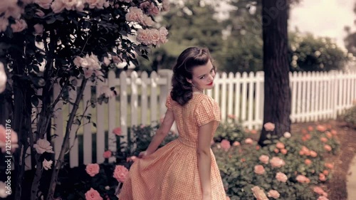 A young woman in a vintage dress poses gracefully in a rose garden beside a white picket fence. This idyllic scene evokes a sense of nostalgia, romance, and classic beauty. Perfect for projects relate