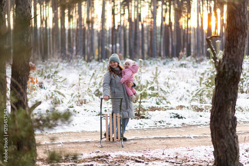 Mother walking in snowy forest with daughter in pink snowsuit and sled photo